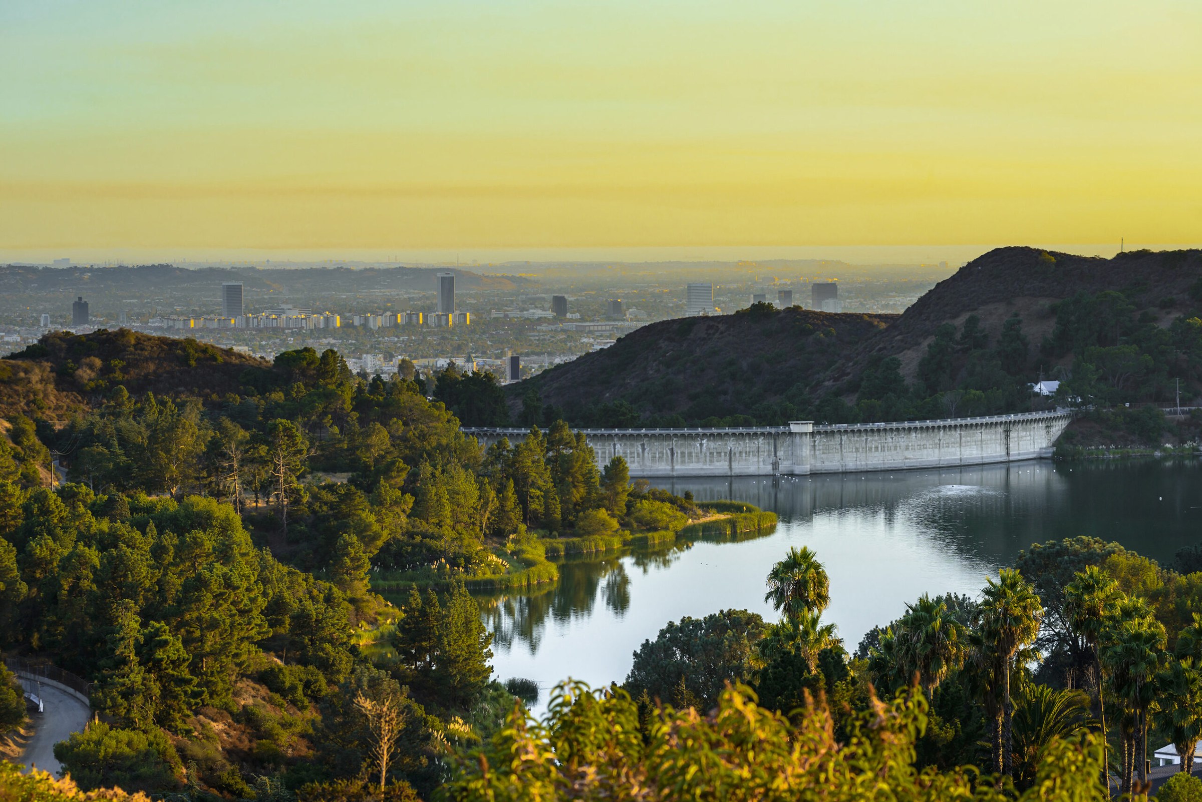 Lake Hollywood and its curved dam at sunset, with the towers of the city far below