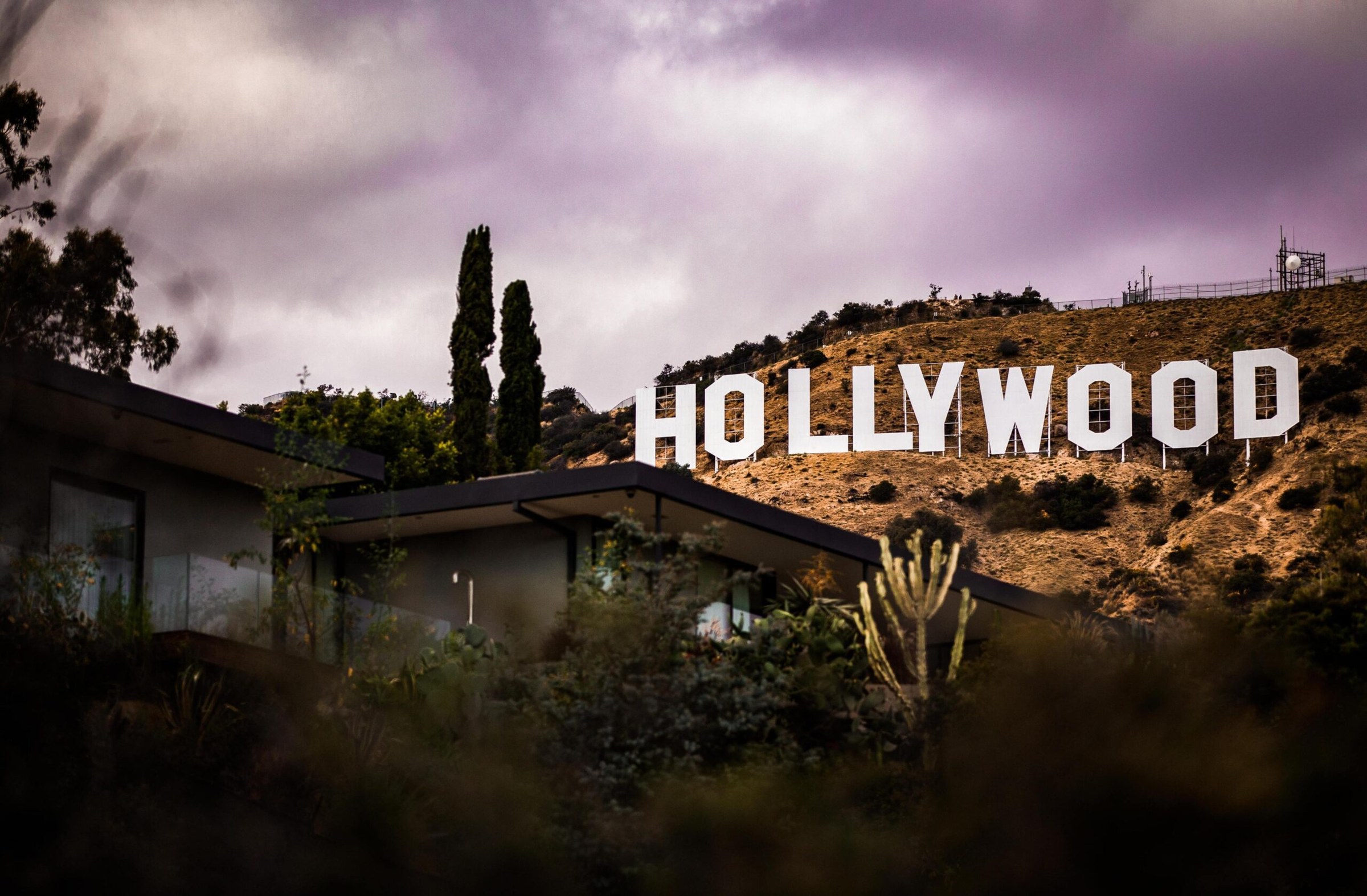 A low modernist house among cacti and trees, with the Hollywood Sign on the hillside behind it under a purple sky